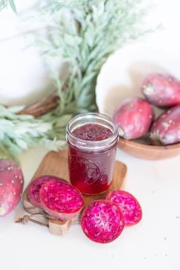 Open jar of prickly pear jelly with sliced fruit showing vibrant magenta interior on a cream background