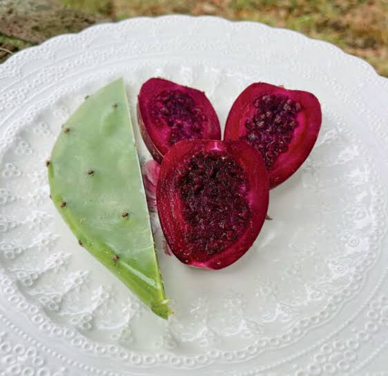 Halved prickly pear fruits showing vivid magenta flesh alongside a green cactus pad on a white plate
